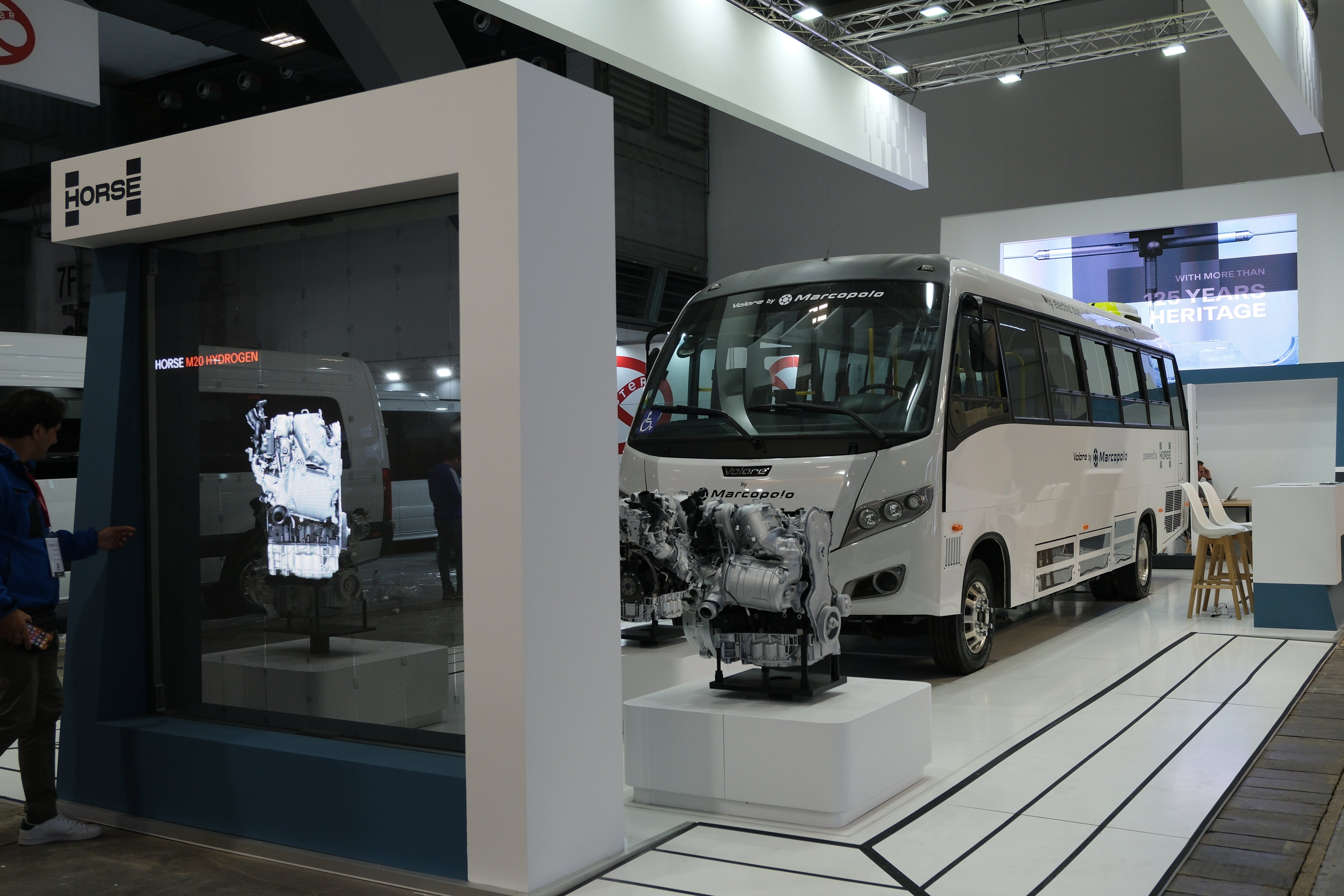 A bus branded with Marcopolo and Horse Powertrain parked at an event exhibit, in front of a screen and behind an engine on display