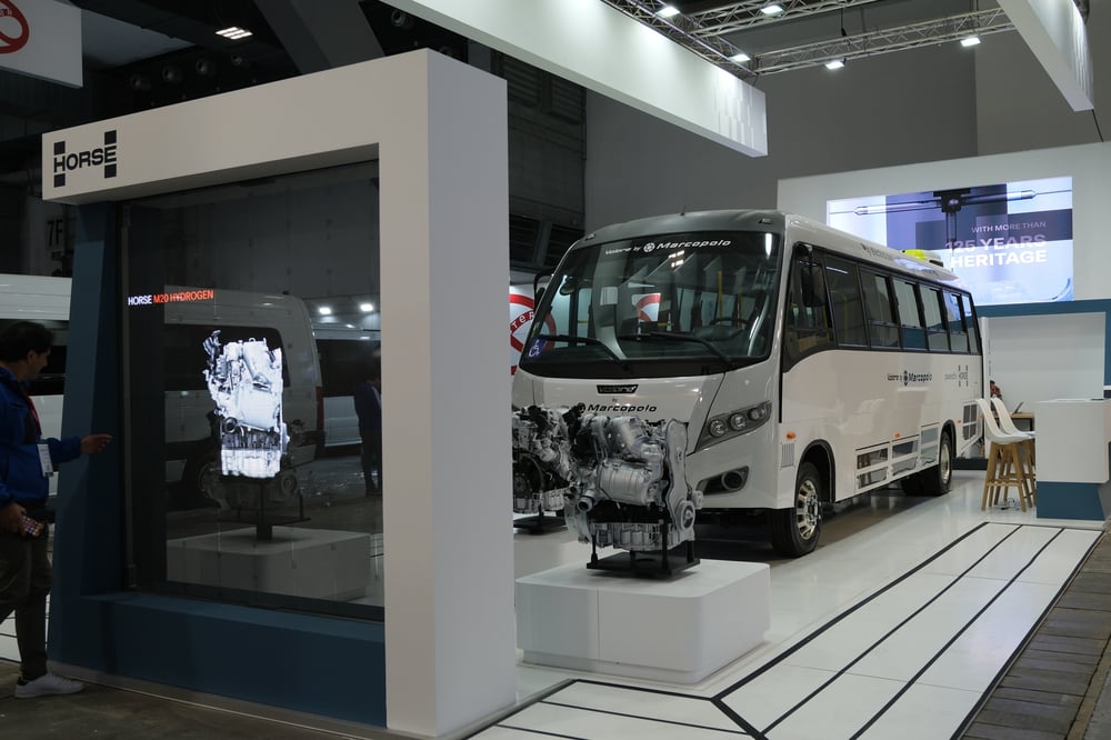 A bus branded with Marcopolo and Horse Powertrain parked at an event exhibit, in front of a screen and behind an engine on display