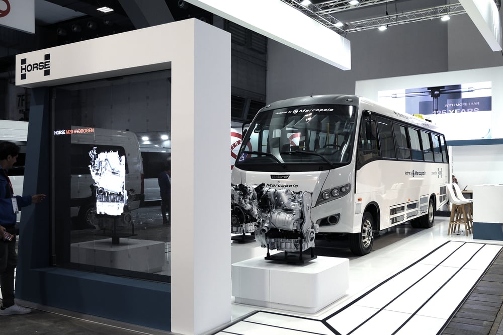 A bus branded with Marcopolo and Horse Powertrain parked at an event exhibit, in front of a screen and behind an engine on display