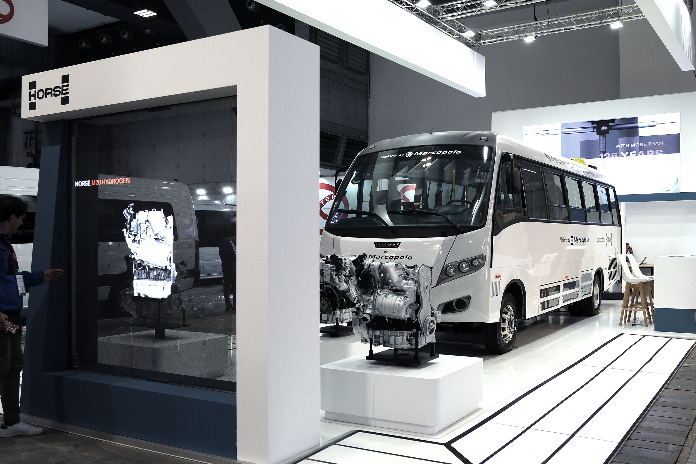 A bus branded with Marcopolo and Horse Powertrain parked at an event exhibit, in front of a screen and behind an engine on display