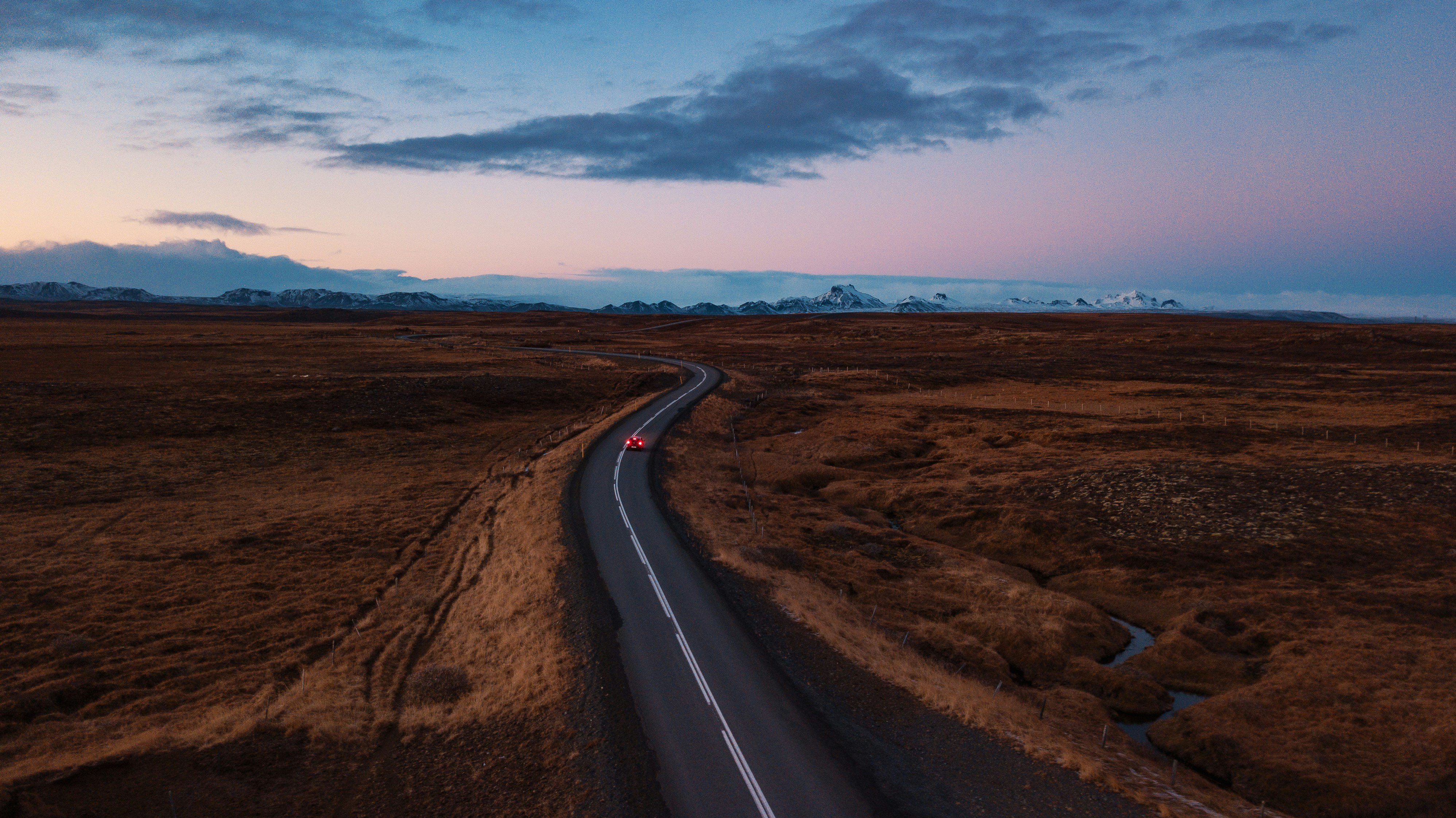An aerial view of a car driving in a treeless, grassy flatland at sunset or sunrise with snow-capped mountains in the distance