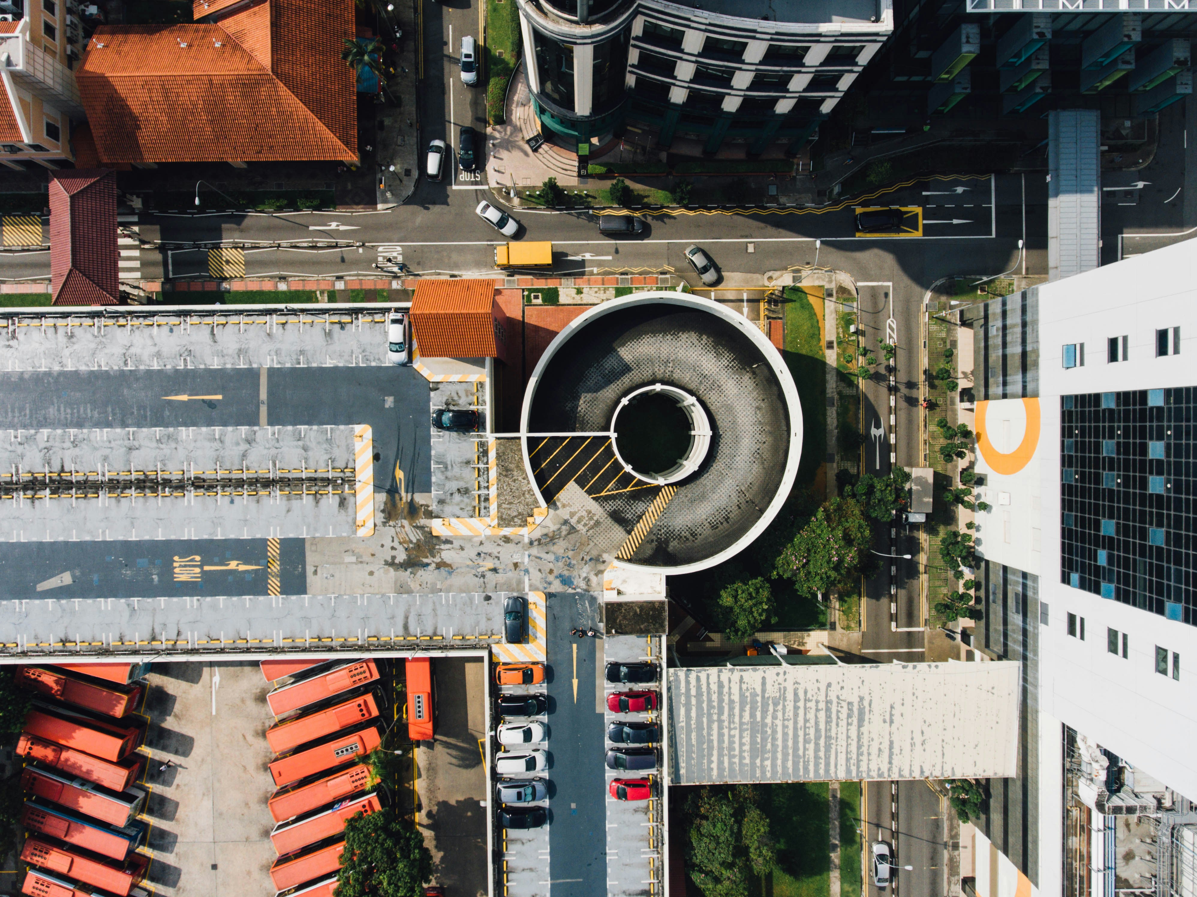 An aerial view of city streets, rooftop car park with cars, and an exposed lower layer with parked buses, with a circular car park entrance at its centre