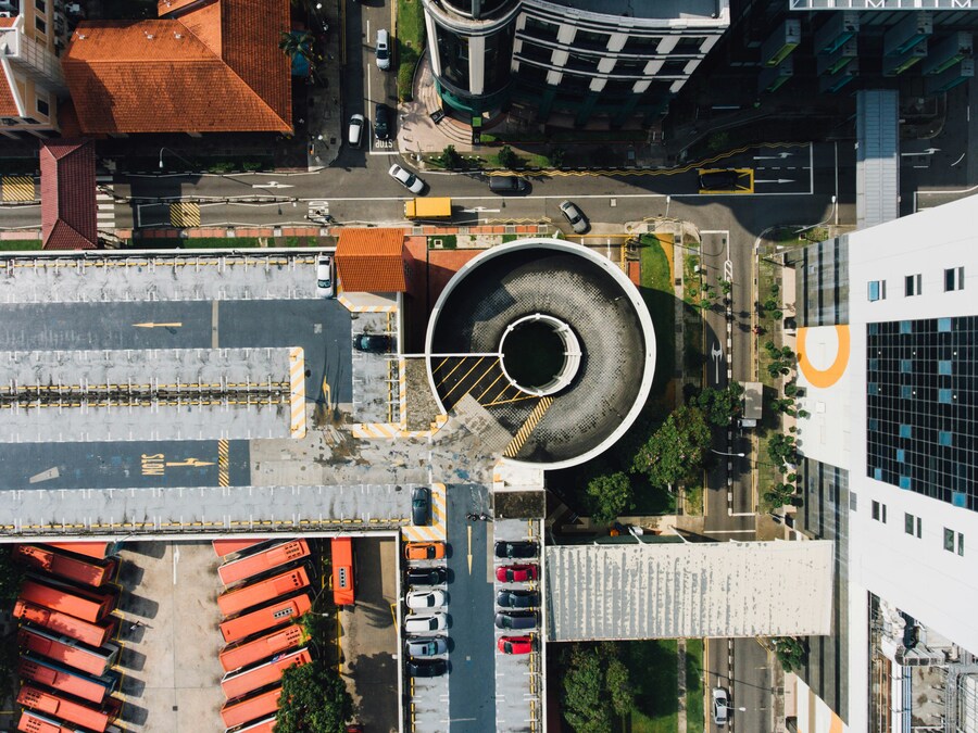 An aerial view of city streets, rooftop car park with cars, and an exposed lower layer with parked buses, with a circular car park entrance at its centre