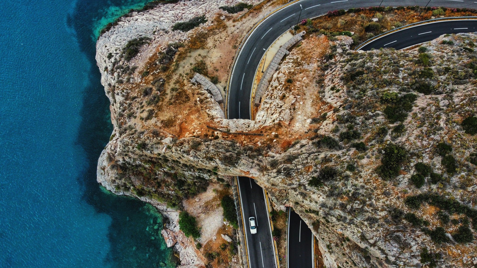 An aerial view of a car driving along cliffs next to clear blue water