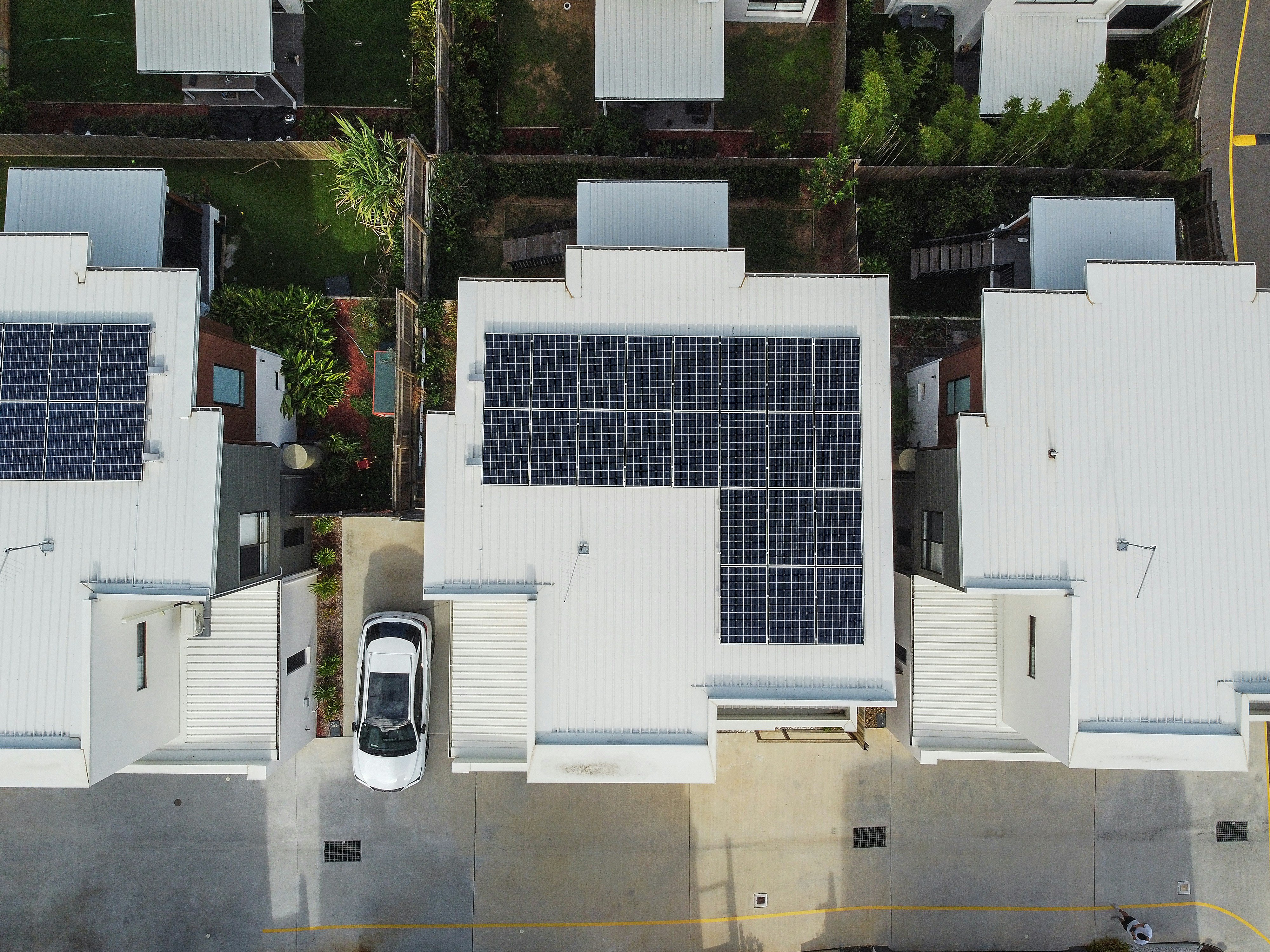 An aerial view of a row of three white houses, two of which topped with solar panels, and the central house with a white car parked on the driveway.
