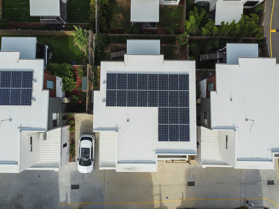 An aerial view of a row of three white houses, two of which topped with solar panels, and the central house with a white car parked on the driveway.