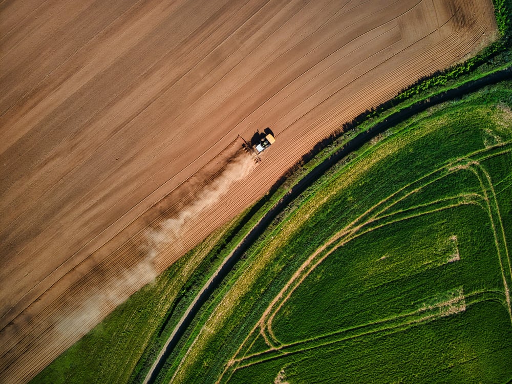 An aerial view of an agricultural vehicle harvesting a brown-coloured field, next to a green-coloured field